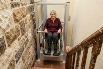 Élévateur PMR vertical installé dans un escalier étroit d'un immeuble ancien en rénovation, avec une femme en fauteuil roulant utilisant la plateforme